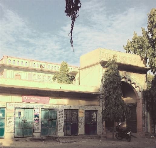 The Shivala temple at Koiripur, the row of shuttered shopfronts in front of it, and a hand-painted Devanagari signboard. It is the first and so far the only street-level photograph of Koiripur found in any open collection. The resolution is low and a filter has been applied; neither has been corrected, because the archive keeps an item as it was found.